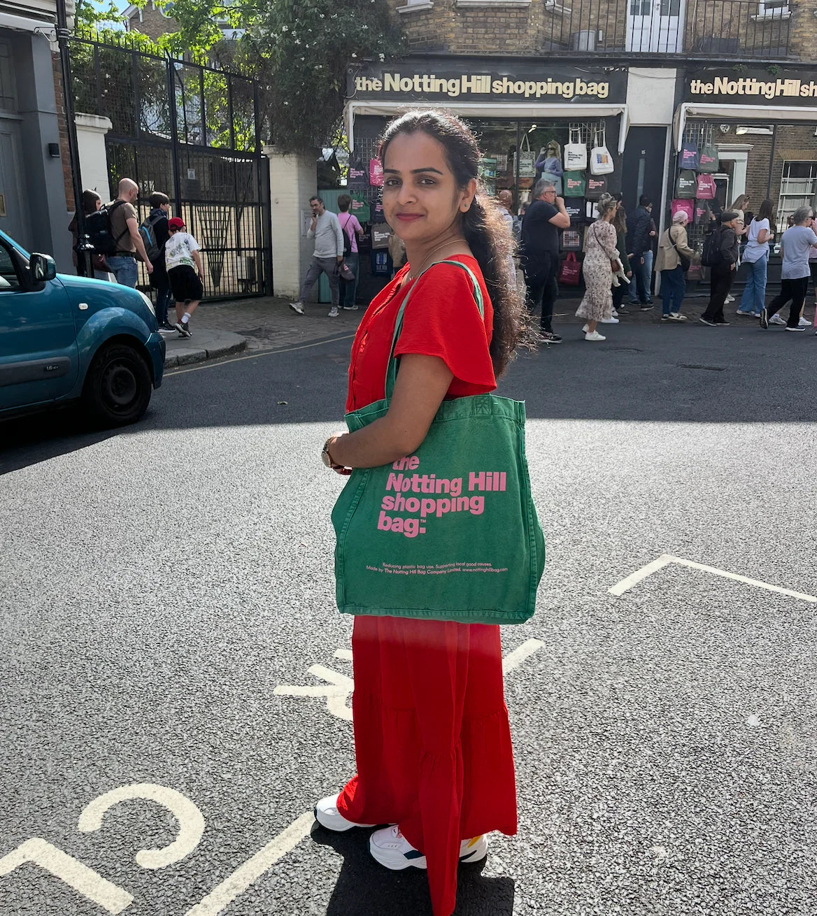A woman in front of the Notting Hill Shop, holding a hand dye collection Notting Hill Shopping Bag, in green with Pink logo
